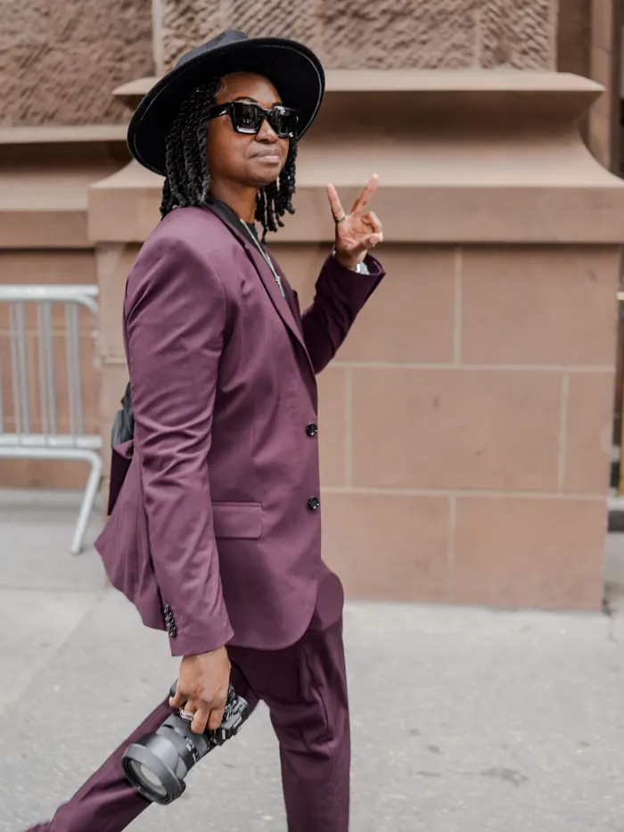 Kash in sunglasses flashing a peace sign as she walks past a brownstone
